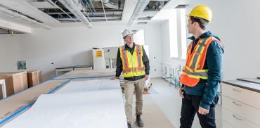 Two construction workers stand six feet apart on an indoor construction site