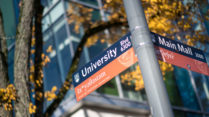 A close-up photo showing a brown and blue Musqueam street sign. This one reads University Blvd and Main Mall with the Musqueam language underneath.