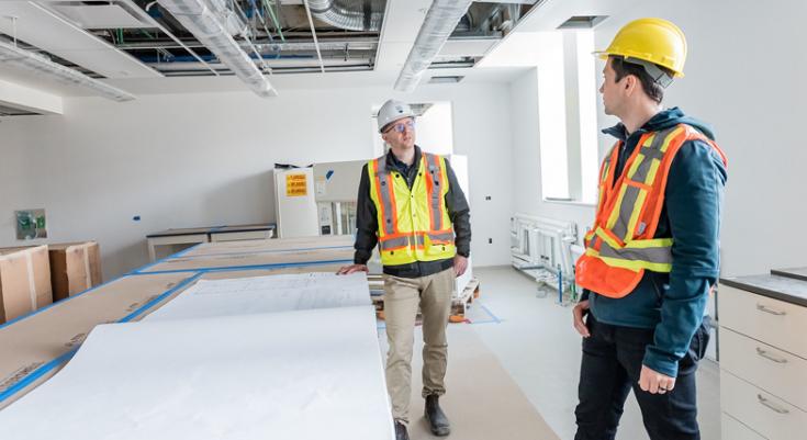 Two construction workers stand six feet apart on an indoor construction site